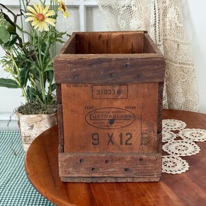 May include: Vintage wooden crate with the text "American Window LUSTRAGLASS" and "9 X 12" printed on the front. The crate is rectangular with a dark brown exterior and metal rivets. The crate is sitting on a wooden surface.