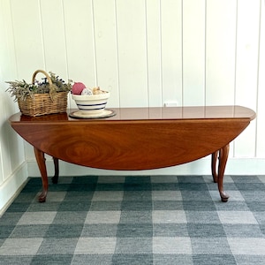 May include: A mahogany drop-leaf table with a smooth, polished surface and curved legs. On top sits a rectangular woven wicker basket filled with dried flowers and a white ceramic bowl with blue horizontal stripes containing decorative spheres.