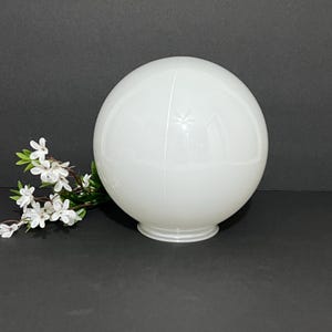 May include: A white, spherical glass globe with a small base. The globe is divided into two halves and has a seam running down the middle. A sprig of white flowers with green leaves rests to the left of the globe.