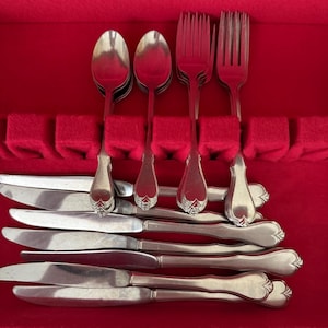 Puede incluir: Un juego de cubiertos antiguos, que incluye cucharas, tenedores y cuchillos, dispuestos en un estuche forrado de terciopelo rojo. La cubertería tiene un diseño decorativo en el mango. El estuche está abierto, revelando los utensilios bien organizados.