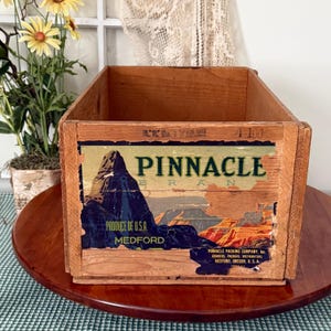 May include: A vintage wooden crate with a colourful label displaying the text "PINNACLE BRAND" and a mountain scene. The label also includes "Produce of U.S.A. Medford" and the Pinnacle Packing Company information. The crate is made of light-coloured wood.
