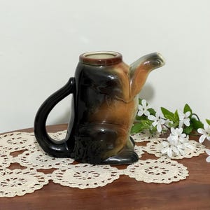 May include: A brown and black ceramic teapot shaped like a dog. The teapot has a spout and a handle. The dog's tail is curled around the handle.