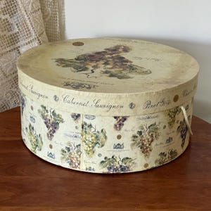 May include: A round, vintage-style box with a lid. The box is decorated with illustrations of grapes and wine-related text. The box has a cream-colored background with a beige lid. A cream-colored rope handle is attached to the side.