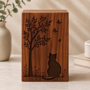 Puede incluir: Urna de madera para mascotas con acabado marrón oscuro. La parte frontal presenta un diseño grabado de un árbol, mariposas, una flor y una silueta de gato. La urna es rectangular y se mantiene en posición vertical.