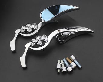 May include: A pair of chrome-plated motorcycle side mirrors featuring a sculpted arm design with three integrated skull motifs. The mirrors have a teardrop-shaped reflective surface and include a set of six threaded metal mounting bolts.