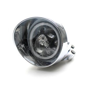 Może przedstawiać: Chromowany reflektor motocyklowy z motywem czaszki wewnątrz soczewki. Reflektor ma okrągły kształt i błyszczącą, odblaskową powierzchnię. Czaszka jest widoczna na środku soczewki.