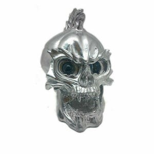 May include: A metallic silver skull figurine featuring a stylized mohawk crest on top. The skull has deep-set dark eye sockets, a prominent brow ridge with leaf-like detailing, and an open jaw revealing teeth, all finished in a reflective chrome.
