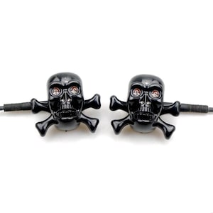 May include: A pair of black skull-shaped turn signal lights with crossbones extending from the sides. Each skull features two clear, circular lenses for eyes and a threaded metal mounting bolt extending from the back for installation purposes.
