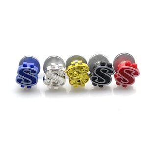 Puede incluir: Cinco pendientes de botón con un diseño de signo de dólar. Los pendientes son de varios colores: azul, plateado, dorado, negro y rojo. Cada pendiente tiene un poste plateado y un emblema de signo de dólar colorido.
