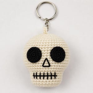Puede incluir: Un llavero de calavera tejido a ganchillo con cuerpo de hilo color blanco hueso, dos ojos circulares de hilo negro, una pequeña nariz triangular negra y una línea horizontal negra bordada como boca, unido a una anilla de metal plateado.