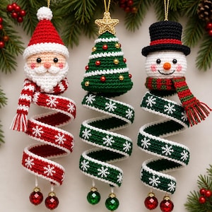 May include: Three crocheted Christmas ornaments featuring a Santa Claus, a green Christmas tree with a gold star, and a snowman wearing a black top hat. Each ornament has a long, spiralling tail decorated with white snowflakes and two dangling beads.
