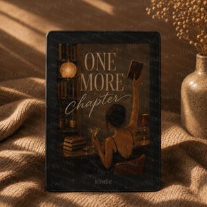 Op de afbeelding: Een zwarte Kindle e-reader met een illustratie van een persoon die een boek omhoog houdt in een bibliotheek met de tekst ONE MORE chapter. Het apparaat heeft een donker frame en een scherm met een warm getinte scène met boekenplanken.