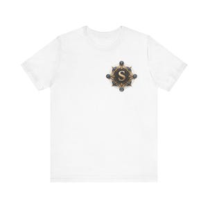 Può includere: T-shirt bianca con un emblema dettagliato sul petto. L'emblema è un design circolare con una combinazione di colori oro e bronzo, e la lettera "S" è ben visibile al centro. Il design include dettagli intricati e un aspetto simile a una bussola.
