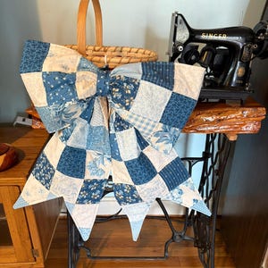 Puede incluir: Un gran lazo decorativo hecho de tela de edredón de patchwork con varios tonos de azul y crema, incluyendo estampados florales, de cuadros vichy y toile. El lazo tiene dos bucles anchos y dos colas largas y puntiagudas que cuelgan.
