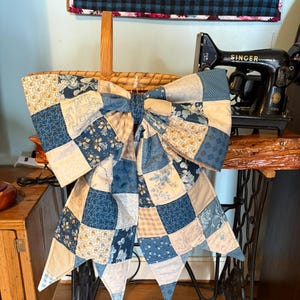 May include: A large decorative bow constructed from a patchwork of quilted fabric squares in various shades of blue, cream, and tan. The fabric features diverse patterns including floral prints, small geometric shapes, and line drawings of houses.