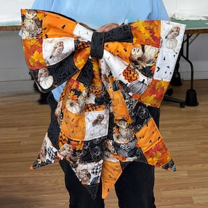 May include: A large, quilted fabric bow featuring a patchwork design with autumn-themed patterns. The fabric displays owls, pumpkins, fall leaves, and black spiderweb motifs in shades of orange, black, white, and yellow, tied with a black centre knot.