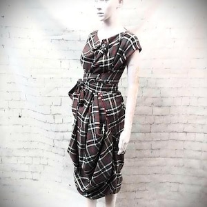 Puede incluir: Vestido a cuadros en tonos burdeos, negro y blanco, exhibido en un maniquí. El vestido presenta un escote drapeado, una cintura ceñida con lazo y una falda fruncida. La tela tiene un patrón de tartán clásico.