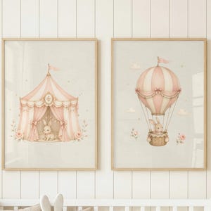 May include: Two framed wall art prints featuring soft pink and cream illustrations. The left print shows a circus tent with a small rabbit inside. The right print depicts a hot air balloon carrying a rabbit in its basket, accented by small flowers.