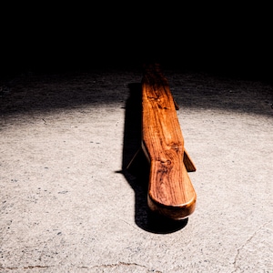 Puede incluir: Banco de madera artesanal con un acabado marrón cálido. El banco presenta un asiento curvo y patas anguladas, mostrando la veta natural de la madera. El diseño es minimalista y moderno, adecuado para uso interior o exterior. Mide unos 150 cm de largo.