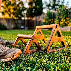 May include: Two wooden parallettes for fitness training, featuring a dark brown finish and a triangular base design. Each piece consists of a horizontal cylindrical bar supported by two triangular side frames, resting on a green grass surface.