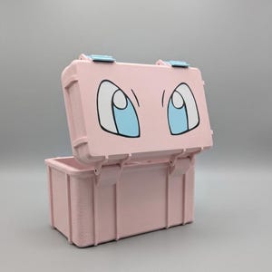 May include: A pink, rectangular storage box with a hinged lid. The lid features a cartoon eye design with black outlines and blue irises. The box is open, revealing the interior.