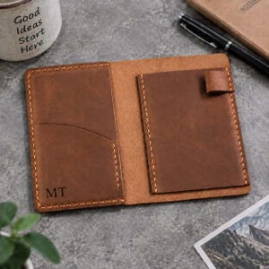 Peut inclure: Un porte-passeport en cuir marron avec des coutures orange visibles, une poche verticale sur le côté gauche avec les initiales MT gravées, une poche rectangulaire sur le côté droit et une petite boucle en cuir pour stylo sur le bord supérieur.