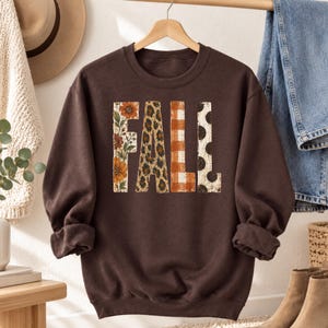 May include: A dark brown crewneck sweatshirt featuring the word FALL printed in large block letters. Each letter has a distinct pattern: the F has orange flowers, the A has a leopard print, the L has orange plaid, and the L has black polka dots.