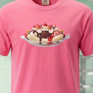 以下が含まれることがあります： 鮮やかなピンク色の半袖Tシャツで、中央にガラス皿に入ったバナナスプリットのグラフィックが描かれています。デザートにはバニラ、チョコレート、ストロベリーのアイスクリームが盛り付けられ、ホイップクリーム、ベリー、チェリーがトッピングされています。