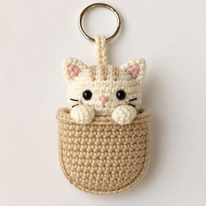 May include: A crochet keychain featuring a cream-colored cat with light brown stripes, pink inner ears, and black eyes, peeking out from a tan crocheted pouch. The cat has two small paws resting on the rim of the pouch, attached to a metal ring.