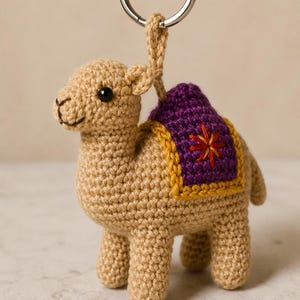 Op de afbeelding: Een beige gehaakte kameel sleutelhanger met een paars zadel met gele rand en een rood en oranje geborduurd sterontwerp. De kameel heeft zwarte kraaloogjes en een kleine gestikte mond, bevestigd aan een zilverkleurige metalen ring.