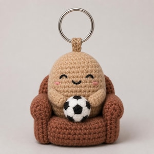 Op de afbeelding: Een gehaakte sleutelhanger met een beige, ovaalvormig figuurtje met een lachend gezicht en roze wangen, zittend in een bruine gehaakte fauteuil terwijl het een kleine zwart-witte voetbal vasthoudt. Een zilveren metalen ring is aan de bovenkant bevestigd.