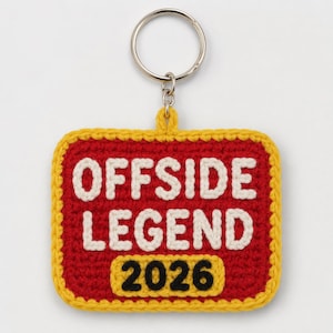 Puede incluir: Un llavero cuadrado de ganchillo con fondo rojo y borde amarillo. El texto "OFFSIDE LEGEND" está bordado en hilo blanco y el número "2026" está bordado en hilo negro dentro de un parche rectangular amarillo en la parte inferior.