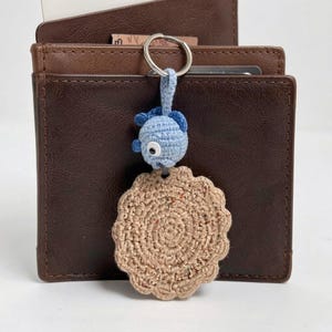 Può includere: Un portafoglio bifold in pelle marrone con un portachiavi all'uncinetto attaccato a un anello in metallo argentato. Il portachiavi presenta un pesce all'uncinetto azzurro con un occhio bianco e nero, collegato a un disco beige festonato.