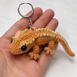 Puede incluir: Un llavero de ganchillo con forma de lagarto con un cuerpo texturizado en color naranja y crema, que presenta grandes ojos de plástico negro y una anilla de metal plateado unida a la parte superior por una cadena corta con puntos detallados.