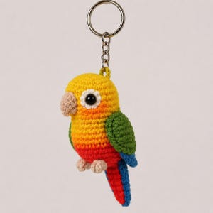 May include: A crochet keychain featuring a small parrot with a yellow head, orange chest, red belly, green wings, and a blue and red tail. The bird has a beige beak, two small beige feet, and a black eye. It is attached to a silver metal keyring.