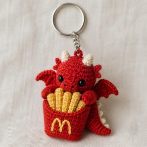 May include: A red crocheted dragon keychain featuring a small red container filled with yellow crocheted french fries. The container displays a yellow embroidered M logo. The dragon has black eyes, cream-colored horns, and a silver metal ring.