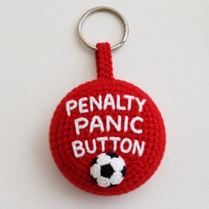 Puede incluir: Llavero rojo tejido a ganchillo con las palabras "PENALTY PANIC BUTTON" en blanco. En la parte inferior aparece un pequeño balón de fútbol. El llavero tiene un anillo de metal plateado.