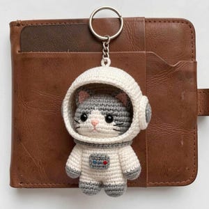 Puede incluir: Un llavero de ganchillo con forma de gato gris y blanco que lleva un casco y traje de astronauta blancos. El gato tiene ojos negros, nariz rosa y bigotes negros. En el pecho tiene un pequeño panel de control rectangular con detalles.