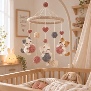 May include: A crochet baby mobile featuring three kittens, each holding a yarn ball. The mobile includes hanging yarn balls in cream, pink, and gray, along with small pink hearts suspended from a circular beige frame with white strings.