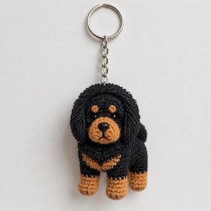 Puede incluir: Un llavero de ganchillo con forma de perro pequeño de color negro y marrón, con orejas caídas, hocico redondeado y ojos oscuros. El perro está unido a una anilla de metal plateado mediante una cadena corta, mostrando un tejido detallado.