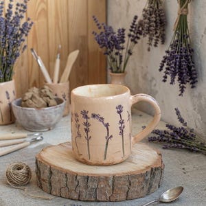 Puede incluir: Una taza de cerámica de color melocotón claro con un patrón de ramas de lavanda prensadas a mano en color morado oscuro. La taza tiene un asa resistente y un acabado esmaltado suave, mostrando un diseño botánico en la superficie exterior.