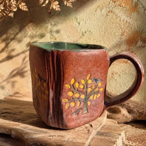 Puede incluir: Una taza de cerámica de forma cuadrada con un exterior texturizado de color marrón rojizo y un interior verde liso. La taza presenta un diseño pintado a mano de un árbol con hojas amarillas y pequeños pájaros negros en los lados y un asa curva.