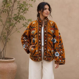 Pode incluir: Uma jaqueta de veludo amarelo mostarda com bordados florais intrincados em tons de azul, vermelho e branco. A jaqueta tem gola alta, mangas compridas e bainha reta, usada sobre uma blusa branca e calças creme de pernas largas.