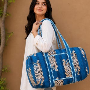 May include: A quilted duffel bag in a deep blue hue featuring a repeating pattern of beige tigers. The bag has two long, striped blue and white fabric handles and a cylindrical shape with stitched detailing throughout the exterior fabric.