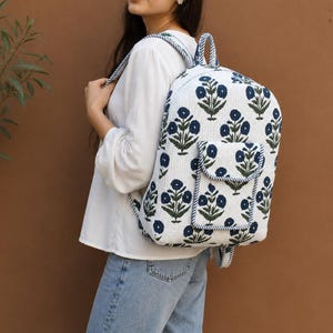 Puede incluir: Una mochila de tela blanca con un patrón floral repetido en azul oscuro. La bolsa incluye un asa superior, correas ajustables para los hombros y un bolsillo exterior frontal con cierre de solapa, todo acabado con un borde a rayas azules y blancas.
