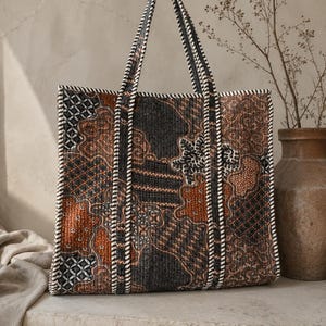 May include: A rectangular tote bag featuring a quilted texture and a dark brown, black, and white batik-style pattern. The bag includes two long shoulder straps and white and black striped piping along the edges and the strap seams.
