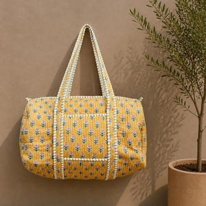 Könnte beinhalten: Gelbe, gesteppte Tragetasche mit einem Blumenmuster in Rosa und Weiß. Die Tasche hat zwei Griffe und eine weiße Bordüre. Die Tasche ist aus einem weichen, gesteppten Material gefertigt. Die Tasche hat eine gute Größe zum Tragen von Gegenständen.