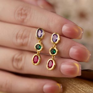 May include: A pair of gold-toned drop earrings featuring three vertically aligned oval-shaped stones in each piece. The top stone is purple, the middle stone is green, and the bottom stone is red. Each gemstone is set in a textured gold-colored metal frame.