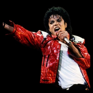 May include: A person wearing a red sequined jacket with white accents and a white shirt, singing into a microphone. The jacket has black trim and is brightly lit against a black background.