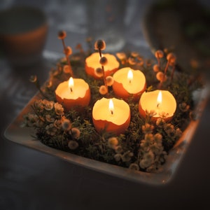 Peut inclure: Un plat en céramique blanche rempli de mousse verte et de fleurs blanches, avec cinq bougies allumées dans des coquilles d'œufs.